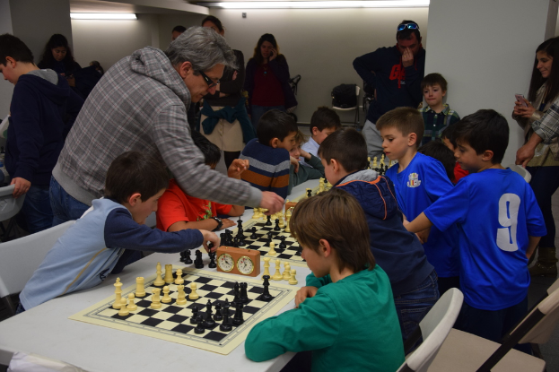 Kinder spielen Schach in einem Wettbewerb, während Erwachsene aus dem Hintergrund und den Seiten zusehen.