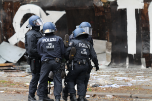 Polizeibeamte in schwarzen Uniformen und blauen Helmen stehen vor einem mit Graffiti bedeckten Gebäude während einer Unruhen, mit verstreuten Papieren und Gegenständen auf dem Boden.
