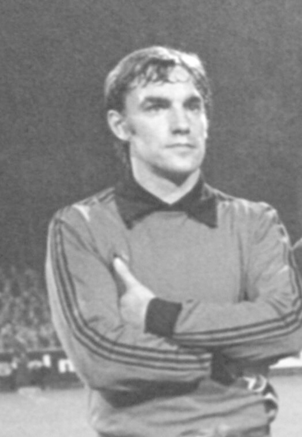 Schwarzes und weisses Foto eines Mannes, der auf einem Fussballfeld steht und die Arme verschraubt hat, umgeben von sitzenden Zuschauern.