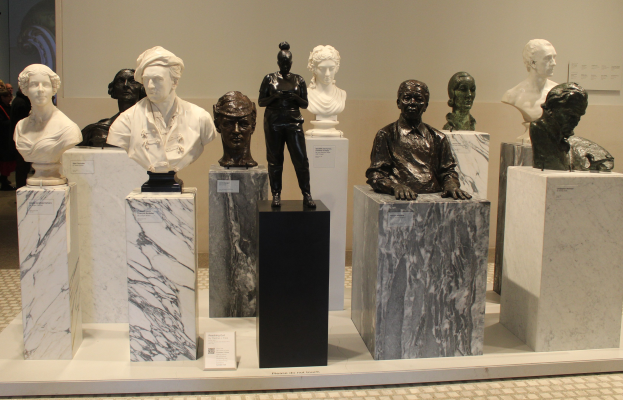 Gruppe von lebensgroßen Büsten-Skulpturen, die in einem Museum ausgestellt sind, mit Besuchern im Hintergrund und einer Wand dahinter.