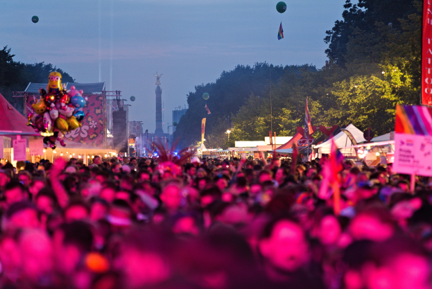 Eine große Menschenmenge auf einem nächtlichen Musikfestival, beleuchtet von bunten Lichtern und Ballons, mit Zelten, Bannern, Fahnen und anderen Gegenständen, die herumliegen, Bäumen, einem Turm und einem sternenklaren Himmel im Hintergrund.