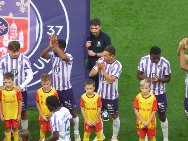 Eine Gruppe junger Jungs auf einem Fußballfeld, die klatschen, mit einem Banner mit Text und Logos im Hintergrund auf saftig grünem Gras.