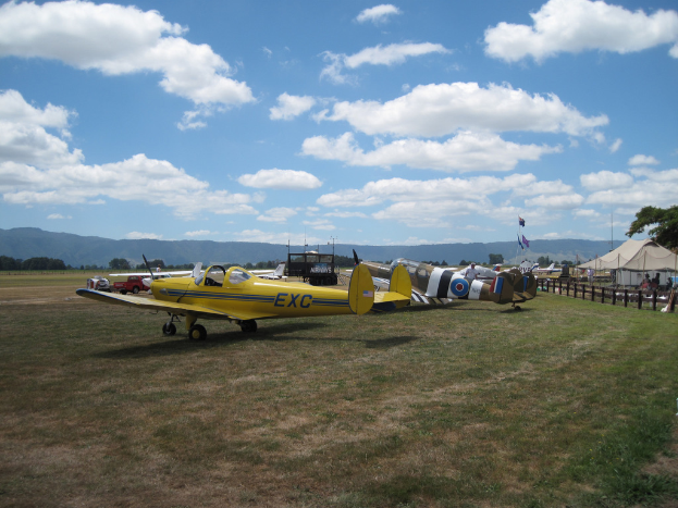 Flugzeuge, Fahrzeuge, Zelte und Fahnen mit Stangen sind auf einem grasbewachsenen Bereich mit Bäumen und Hügeln im Hintergrund unter einem klaren Himmel aufgebaut.