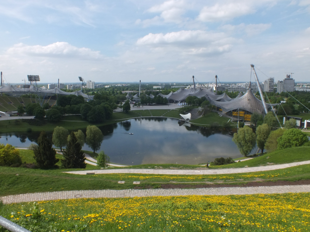 Blick auf das Olympische Stadion von einem Hügel aus, mit einem Teich im Vordergrund, umgeben von Grün und gelben Blumen, und Gebäuden im Hintergrund unter einem klaren blauen Himmel.