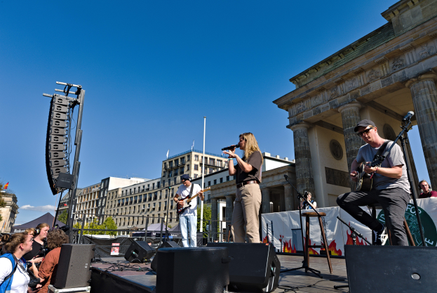 Eine Gruppe von Menschen auf der Bühne vor dem Brandenburger Tor in Berlin, Deutschland, die Musikinstrumente spielen und in Mikrofone singen, mit Lautsprechern und anderen Gegenständen drumherum, und Gebäuden, Bäumen, Pfählen, Flaggen und einem klaren blauen Himmel im Hintergrund.