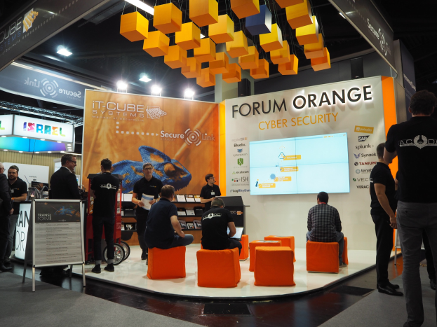 Eine Gruppe von Menschen an einem IT-Cube Systems Forum Orange Cyber Security Stand, mit sitzenden und stehenden Teilnehmern, einem Anzeigebildschirm, Texttafeln, Regalen mit Gegenständen, Deckenleuchten und Lampen.