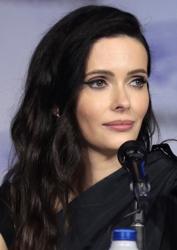 Krysten Ritter auf der Comic-Con in San Diego, in einem schwarzen Kleid, vor einem Mikrofon mit einer Flasche unten, mit einem unscharfen, traumhaften Hintergrund.