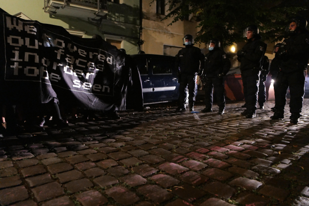 Gruppe von Polizisten mit Helmen, die ein Banner auf einem Kopfsteinpflasterweg bei Nacht halten, mit Fahrzeugen, Gebäuden, Bäumen und Straßenbeleuchtung im Hintergrund.
