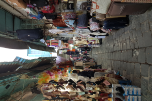 Ein belebter Straßenmarkt mit verschiedenen Artikeln wie Taschen, Kleidung und Schuhen, die zum Verkauf angeboten werden, mit Menschen, die durch die Gänge schlendern und einer Wand auf der rechten Seite, Gebäuden und einem klaren blauen Himmel im Hintergrund.