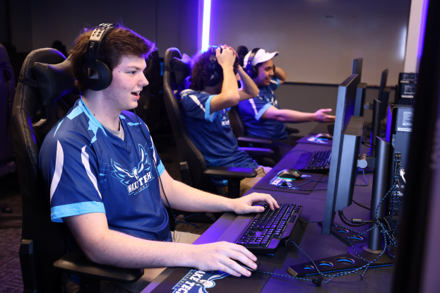 Gruppe junger M├Ąnner mit Headsets, die ein Videospiel auf Computern mit Monitoren, Tastaturen und M├Ąusen an einem Schreibtisch spielen, vor einer beleuchteten Wand.