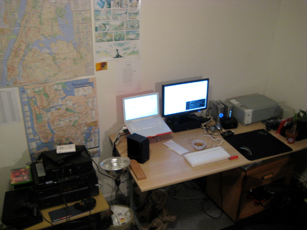 Ein Raum mit einem Tisch, auf dem ein Laptop, ein Lautsprecher, ein Monitor, eine Tastatur, ein Ball, eine Maus, ein Scanner und Papiere liegen, und ein weiterer Tisch mit Elektronikgeräten, sowie Charts und Poster an der Wand dahinter.