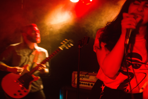 Eine Frau singt in ein Mikrofon auf der Bühne, während ein Mann im Hintergrund Gitarre spielt und Deckenlampen das lebendige Ambiente beleuchten.