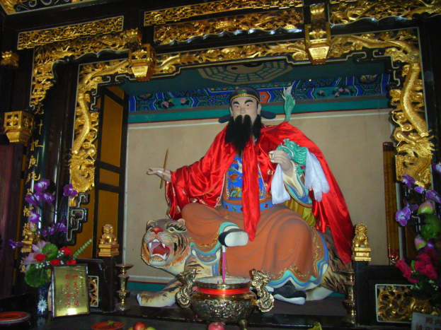 Eine Statue eines chinesischen Mönchs in buntem Gewand sitzt auf einer Tigerstatue innerhalb eines Bogens mit goldenen Verzierungen, vor sich ein Blumenkübel und in der Mitte eine Schüssel.