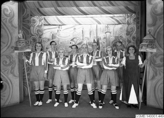 Eine Gruppe strahlender Frauen in einem 1940er-Fußballteam posiert auf einer Bühne, jede hält einen Stock in der Hand, mit einer bemalten Wand im Hintergrund.