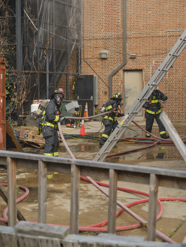 Eine Gruppe von Feuerwehrmännern in Helmen, die Rohre halten, arbeiten daran, ein Gebäude Feuer zu löschen, mit verschiedenen Ausrüstungen und Strukturen drumherum, einschließlich eines Metallzauns, Rohre auf dem Boden, ein Container, ein Verkehrskegel, einige Gegenstände auf der Oberfläche, ein Gebäude mit Fenstern und einer Tür, ein Metallrahmen, Drähte, ein Baum und der Himmel im Hintergrund.