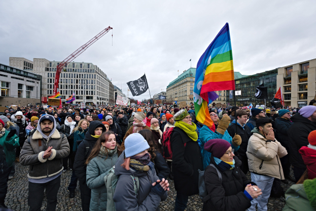 Gro├če Gruppe von Menschen mit LGBTQ+-Rechten-Schildern und -flaggen bei einem Marsch in Berlin, mit Geb├Ąuden, einem Kran und einem bew├Âlktem Himmel im Hintergrund.