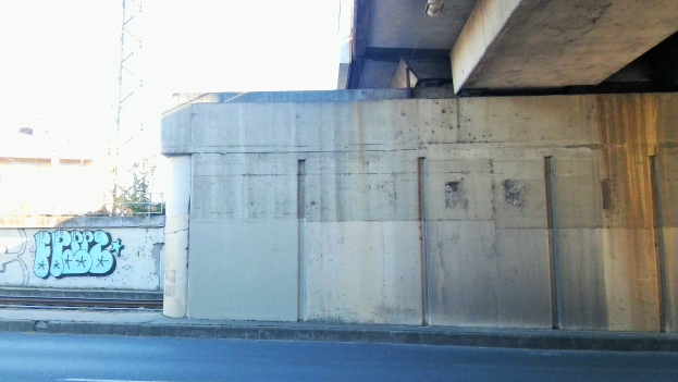 Betonwand mit Graffiti, Straße am Fuß, ein Gebäude und ein Turm im Hintergrund unter einem klaren blauen Himmel.