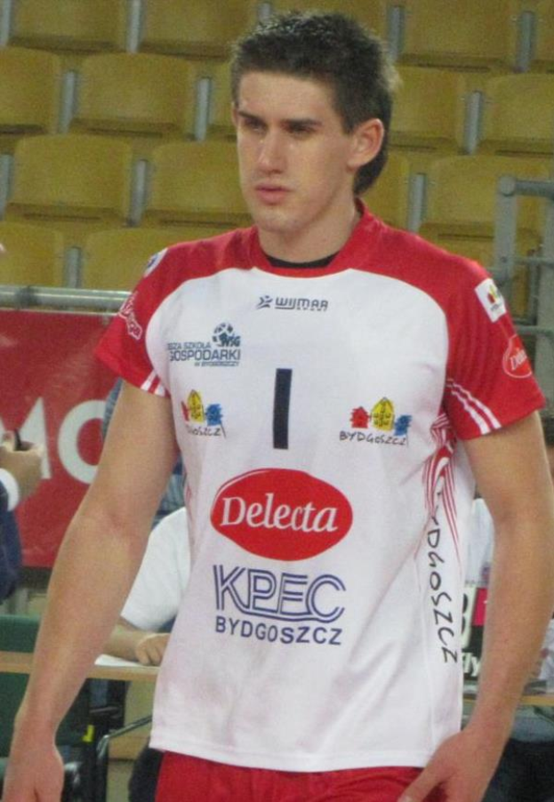 Ein Mann in rot-weißer Handballuniform steht auf einem Feld, umgeben von Zuschauern, mit einem roten Banner im Hintergrund, das "Krasnoyarsk vs Bydgoszcz" trägt und entschlossen wirkt.