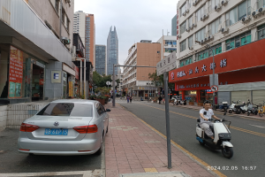 Ein Mann fährt mit einem Roller eine Straße entlang, die von hohen Gebäuden gesäumt ist, mit Menschen auf den Bürgersteigen und Fahrzeugen auf der Straße, unter einem klaren blauen Himmel mit Bäumen im Hintergrund.