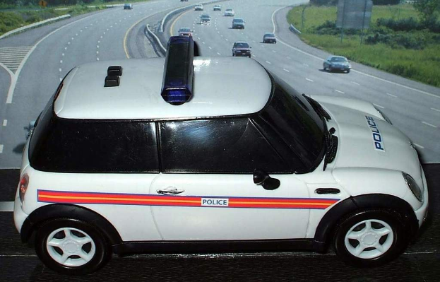 Weißer Polizeiwagen mit "Polizei"-Aufschrift vor einer Reihe fahrender Fahrzeuge auf einer von Gras und Bäumen gesäumten Straße außerhalb der Stadt.