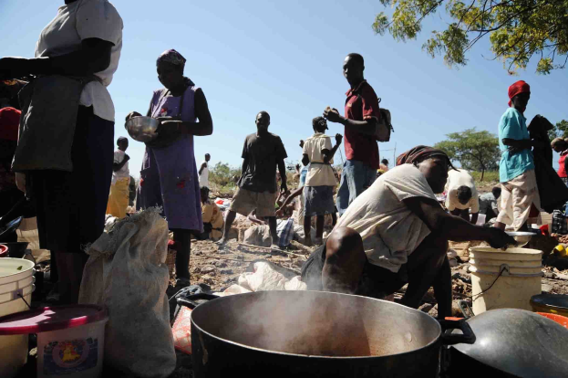 Eine Gruppe von Menschen bereitet Essen im Freien in einer ländlichen Umgebung zu, mit einem großen Topf, Eimern und anderen Gegenständen auf dem Boden, unter einem klaren blauen Himmel.