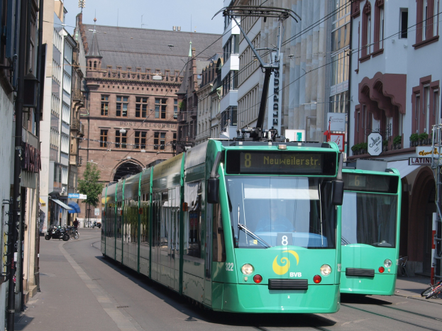 Zwei grüne Straßenbahnen fahren eine von hohen Gebäuden gesäumte Stadtstraße entlang, mit parkenden Fahrrädern und Fußgängern auf den Gehwegen, einem Baum und einem klaren blauen Himmel im Hintergrund.