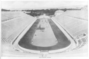 Ein altes Schwarz-Weiß-Foto des Olympischen Stadions in Athen, Griechenland, mit Sitzreihen, Bäumen, Gebäuden und Bergen im Hintergrund sowie Text am unteren Bildrand.