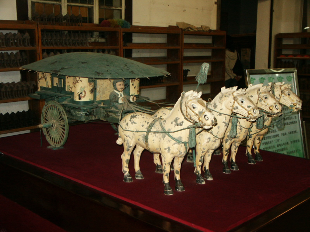 Puppenpferde ziehen einen Wagen auf einem roten Teppich mit einer Person darin, vor Regalen mit Gegenständen und einer Texttafel im Hintergrund.