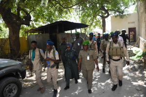 Eine Gruppe von Polizisten in Uniform, einige mit Waffen, die eine Straße entlanggehen, vorbei an einem Auto mit Bäumen, Schuppen, Wänden und Gebäuden im Hintergrund unter einem sichtbaren Himmel.