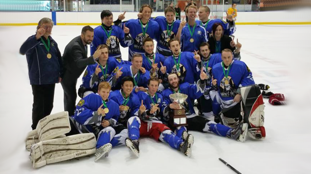 Eine Gruppe von Männern mit Medaillen und einem Pokal auf einem Eisstadion stehend und kniend, feiern ihren Meisterschaftssieg vor Glaswänden und anderen Gegenständen im Hintergrund.