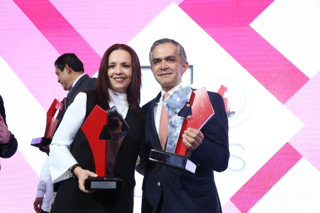 Zwei Menschen lächeln, während sie Trophäen auf einer Bühne halten, mit einem Bildschirm im Hintergrund.