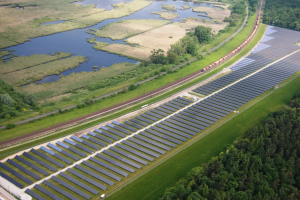 Luftaufnahme einer Solarpark mit Panelen in einem Feld, umgeben von Bäumen, Gras, Pflanzen und Wasser, mit einem Zug auf einem nahen Bahngeleis.