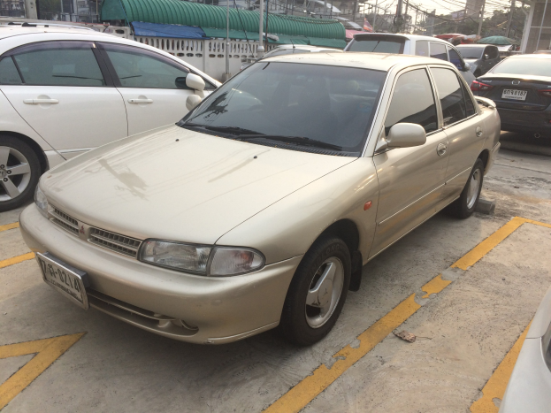 Goldfarbener Wagen auf einem Parkplatz zwischen anderen Fahrzeugen geparkt, mit Gebäuden, Strommasten, Bäumen und einem klaren blauen Himmel im Hintergrund.