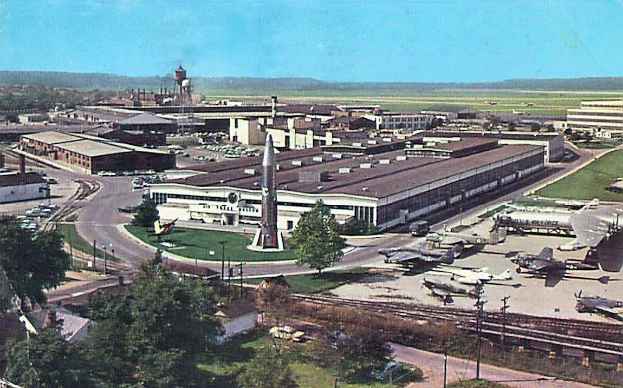 Alte Schwarz-Weiß-Flughafenszene mit geparkten Propellerflugzeugen, Terminalgebäuden, Bäumen, Straßen, Fahrzeugen, Versorgungsmasten, Gras, Bahnschienen, fernen Hügeln und einem klaren Himmel, mit Untertext am unteren Rand.