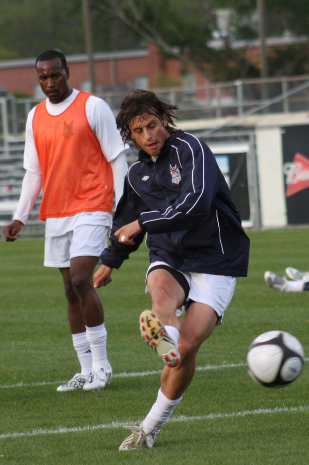Ein Fussballspieler in einer schwarzen Jacke und weissen Shorts kickt einen Ball, mit einem anderen Spieler in einem orangen Jackett dahinter und einem eingezäunten Bereich mit Bäumen im Hintergrund.