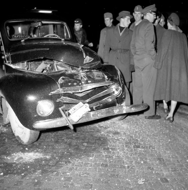 Ein Schwarz-Weiß-Bild eines verunglückten Autos am Straßenrand mit eingedrückter Haube und zersplitterten Fenstern, umgeben von einer Gruppe besorgter Menschen.
