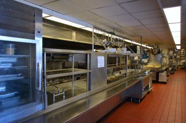 Gewerbeküche mit Edelstahl-Theke, aufgehängten Utensilien, einem Spülbecken, Schränken und diversen Gegenständen, die in Regalen unter elektrischen Deckenleuchten angeordnet sind.