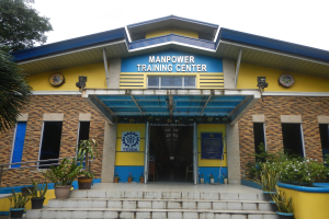 Außenansicht des Manpower Training Centers in Manila, Philippinen, mit Treppen, Topfpflanzen, einem Baum und einem Gebäude mit Fenstern, Türen und Beschriftungen vor einem Himmel-Hintergrund.