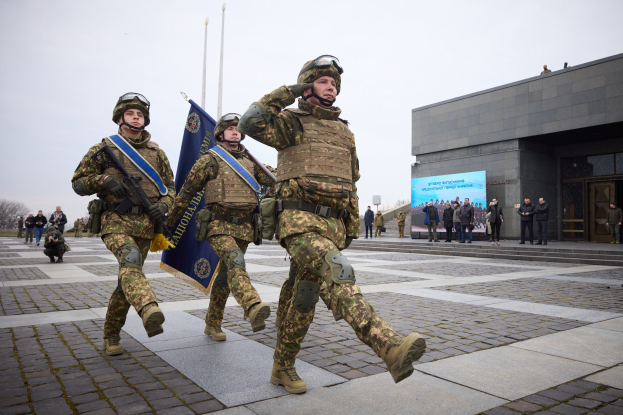 Eine Gruppe ukrainischer Soldaten in Uniform marschiert mit Gewehren und Fahnen die Straße entlang, vor einem Gebäude, Menschen, Bäumen und einem klaren Himmel.