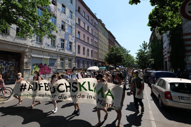 Eine Gruppe von Menschen marschiert mit einem Transparent die Straße entlang, mit parkenden Autos auf der rechten Seite und einem Fahrradfahrer auf der linken Seite, vor Gebäuden und Bäumen in Berlin.
