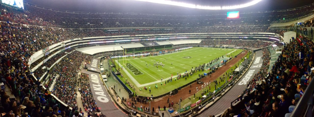 Ein großes Stadion voller Zuschauer, die ein Fußballspiel verfolgen, mit Spielern und verschiedenen Objekten auf dem Feld, beleuchteten Tafeln, Geländern, Bildschirmen und Deckenleuchten.