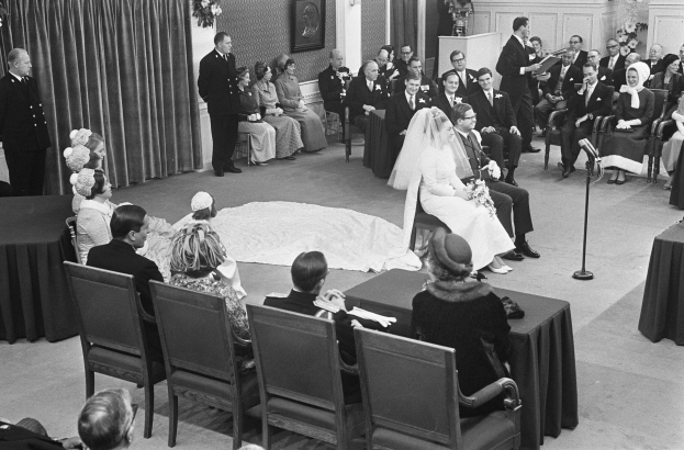 Schwarzes und weißes Foto einer Hochzeitszeremonie mit sitzenden und stehenden Gästen, einem Mann mit Mikrofon, einer Frau mit Blumenstrauß, einem Podium mit Mikrofon auf der rechten Seite, Blumenschmuck und einem Vorhang auf der linken Seite, einem gerahmten Foto und einer Tür im Hintergrund.