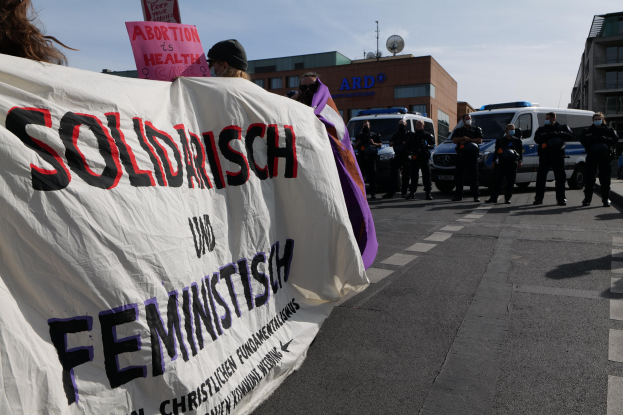 Eine Gruppe von Menschen geht eine Straße entlang und hält eine 'Solidarität und Feminismus'-Schleife hoch, mit parkenden Fahrzeugen, Gebäuden, einer Schüsselantenne und einem klaren blauen Himmel im Hintergrund.