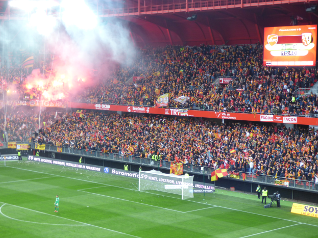 Ein Fußballspiel in einem großen Stadion mit Spielern auf dem Boden, einem Torpfosten, einem Zaun, Bannern, Fahnen, einem Bildschirm, Metallrahmen, einem Dach mit Deckenleuchten und aufsteigendem Rauch.