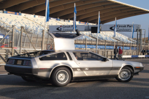 Ein silberner DeLorean DMC-12 auf einer Straße umgeben von Menschen, Fahnen, Schildern, Geländern, Stühlen und einem Schuppen, mit Bäumen und einem klaren blauen Himmel im Hintergrund.