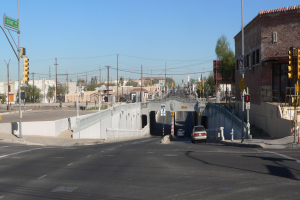 Eine Stadtstraße mit Autos, eine Brücke im Hintergrund, Strommasten mit Drähten, Laternen, Verkehrsampeln, Werbetafeln, Gebäude mit Fenstern, Bäume und ein bewölkter Himmel.