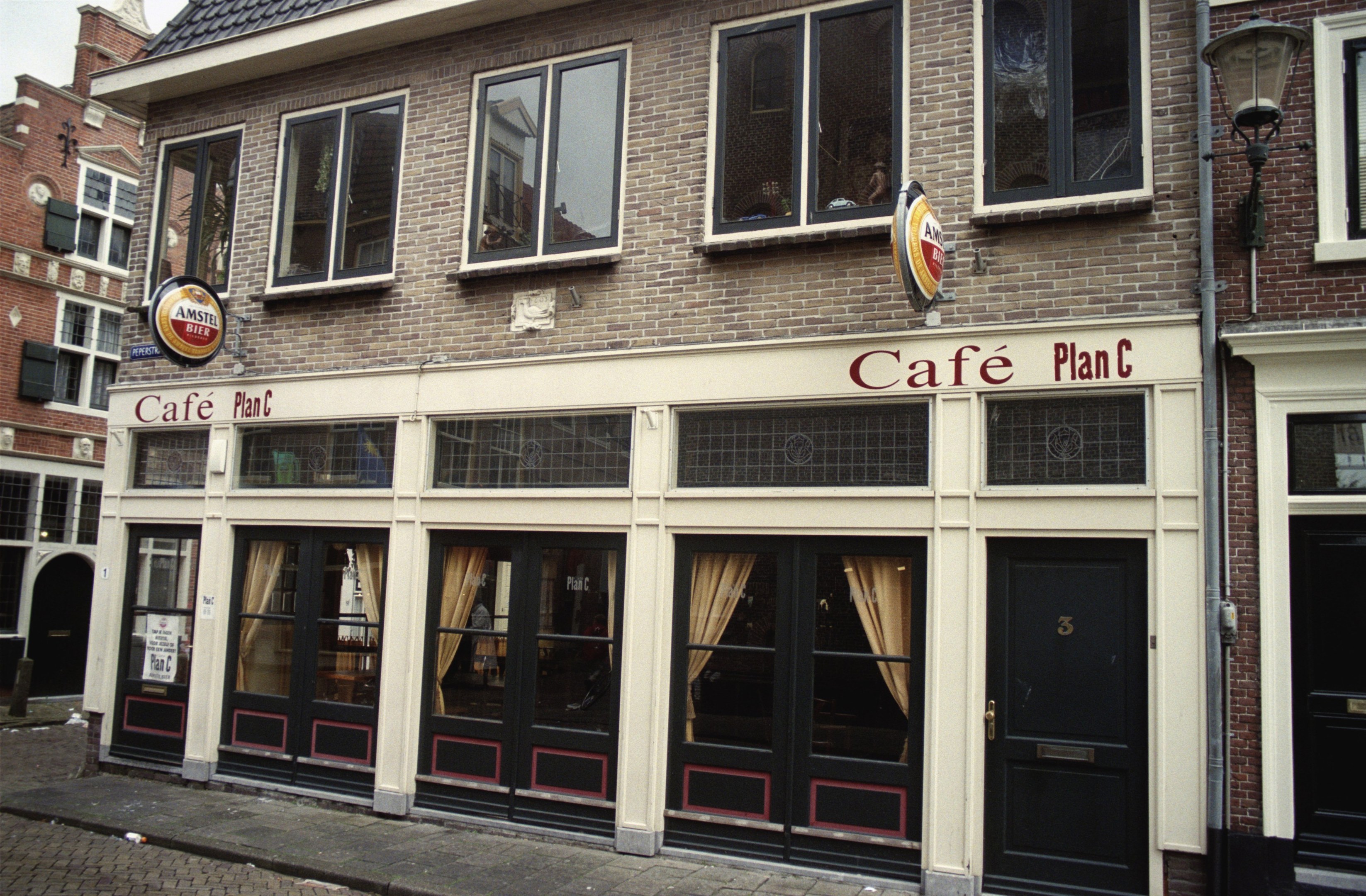 Ein Gebäude mit Fenstern und Türen, ein Schild mit der Aufschrift "Cafe Plan C in Amsterdam, Niederlande", ein Straßenschild, eine Baumgruppe und ein bewölkter Himmel.