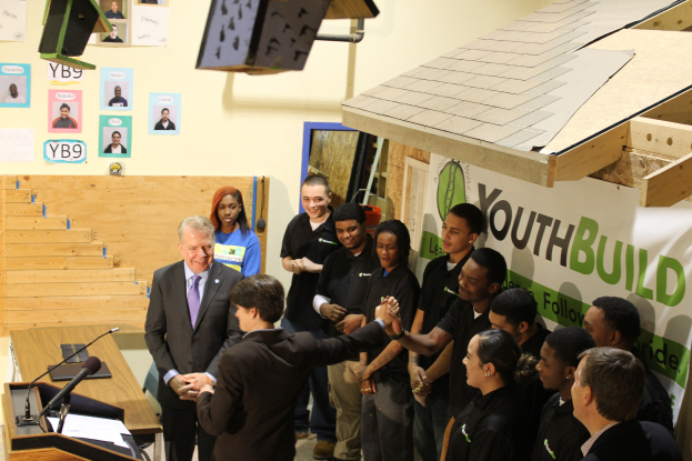 Gruppe von Menschen, die sich die Hände schütteln um ein Podium mit Mikrofonen und Papieren auf einem nahen Tisch, vor einer Menge, mit Plakaten an der Wand und Gegenständen auf dem Dach und rechten Seite.