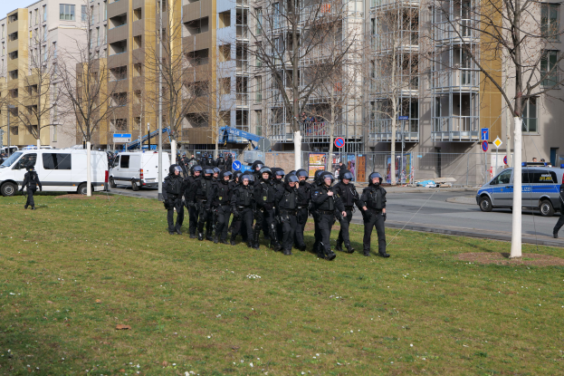 Eine Gruppe von Polizisten in schwarzen Uniformen und Helmen steht auf einem grünen Feld, mit Fahrzeugen auf der Straße davor und Bäumen, Polen, Schildern, Gebäuden und einem klaren blauen Himmel im Hintergrund.