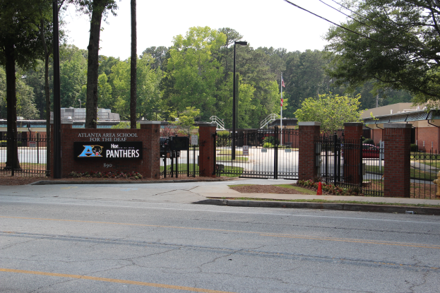 Der Eingang zu einer Schule mit einem Schild an der Seite, umgeben von Bäumen, Straßenlaternen, Gittern, Namensschildern, Fahrzeugen auf der Straße, Absperrgittern, Pflanzen, Gras, Gebäuden und einem klaren blauen Himmel.